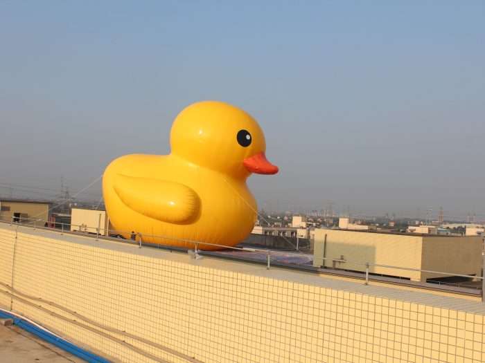 Full side-body view of a giant inflatable yellow rubber duck against a blue sky with a cityscape in the background