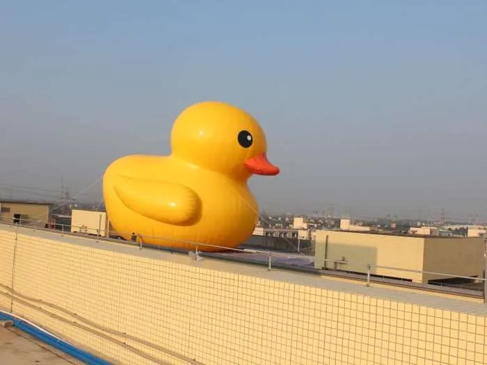Full side-body view of a giant inflatable yellow rubber duck against a blue sky with a cityscape in the background