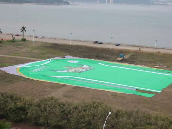 Workers unfurling a giant green PVC jersey panel across an open field lined with palm trees