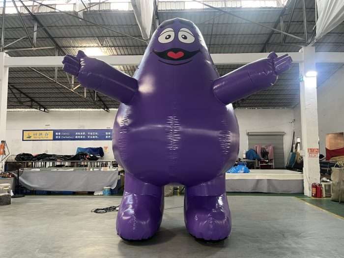 Giant inflatable purple blob-shaped mascot character with open arms and cartoon face, displayed in a warehouse